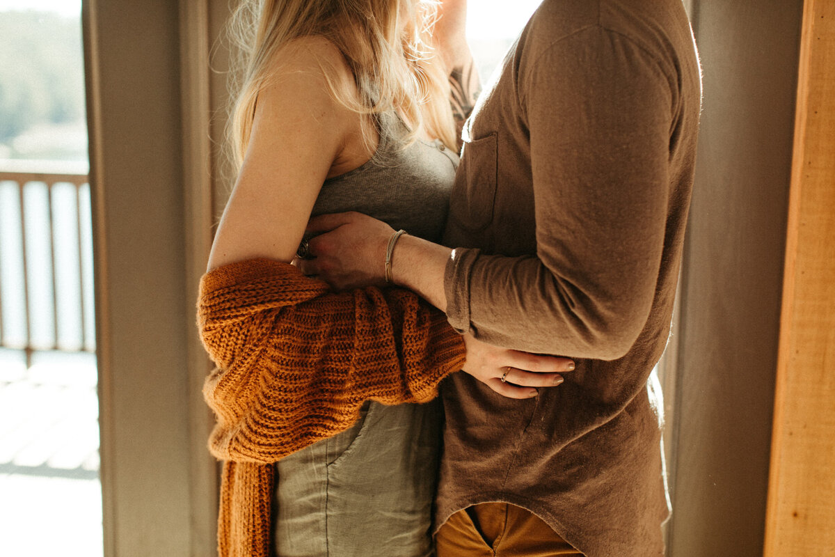 A girl is wearing a loose baggy cardigan around her arms while she holds onto her fiancé in front of a window inside a cabin,