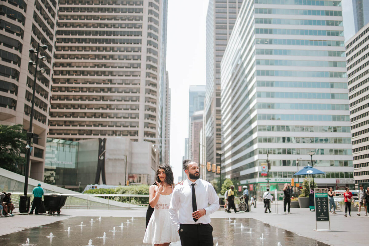 A stylish couple poses confidently in Dillworth Park, surrounded by tall buildings and a water fountain.