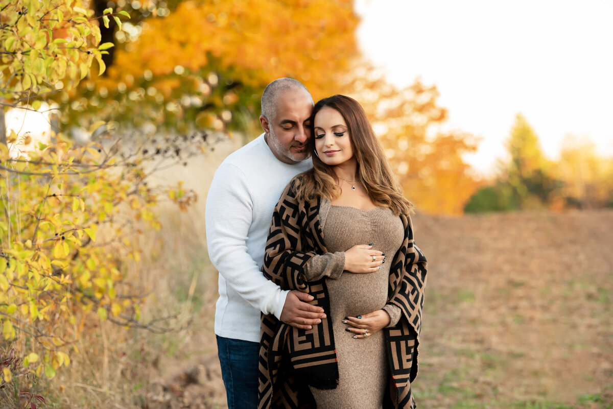 husband and wife standing snuggling cradling her baby belly for her fall outdoor Toronto maternity photography session.
