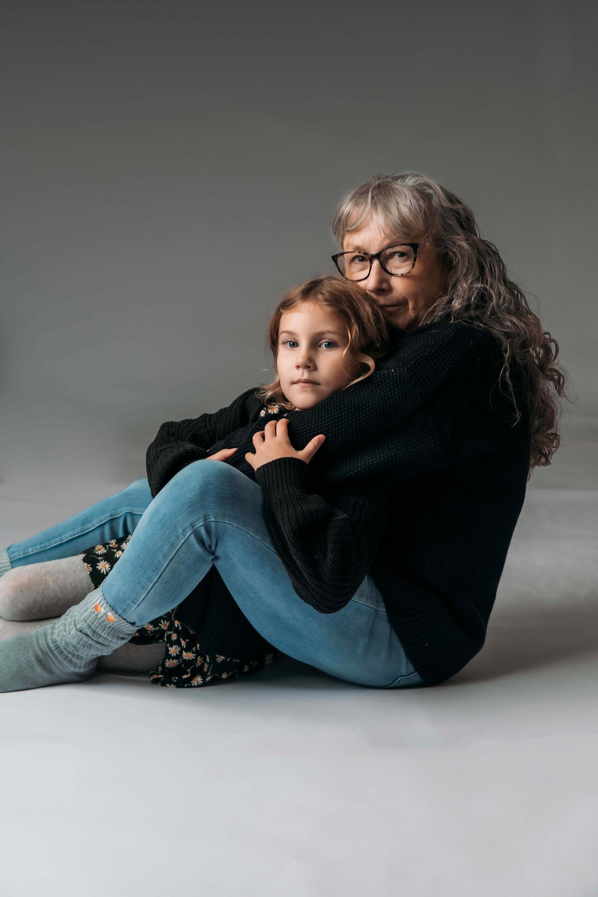 A grandmother wearing a cozy outfit of blue jeans, black sweater, and grey socks sits in the floor of Mobile, AL Motherhood Photographer studio as she pulls in her 6-year old granddaughter for an intimate portrait.