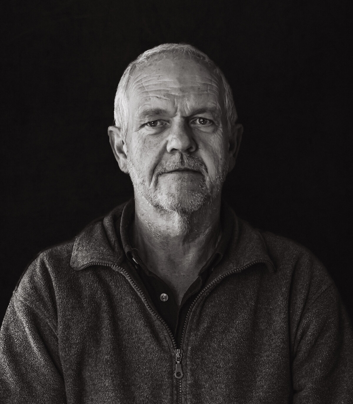 Een zwart wit portretfoto van een oudere man met een vriendelijke uitstraling met glimlach, gefotografeerd tegen een donkere achtergrond