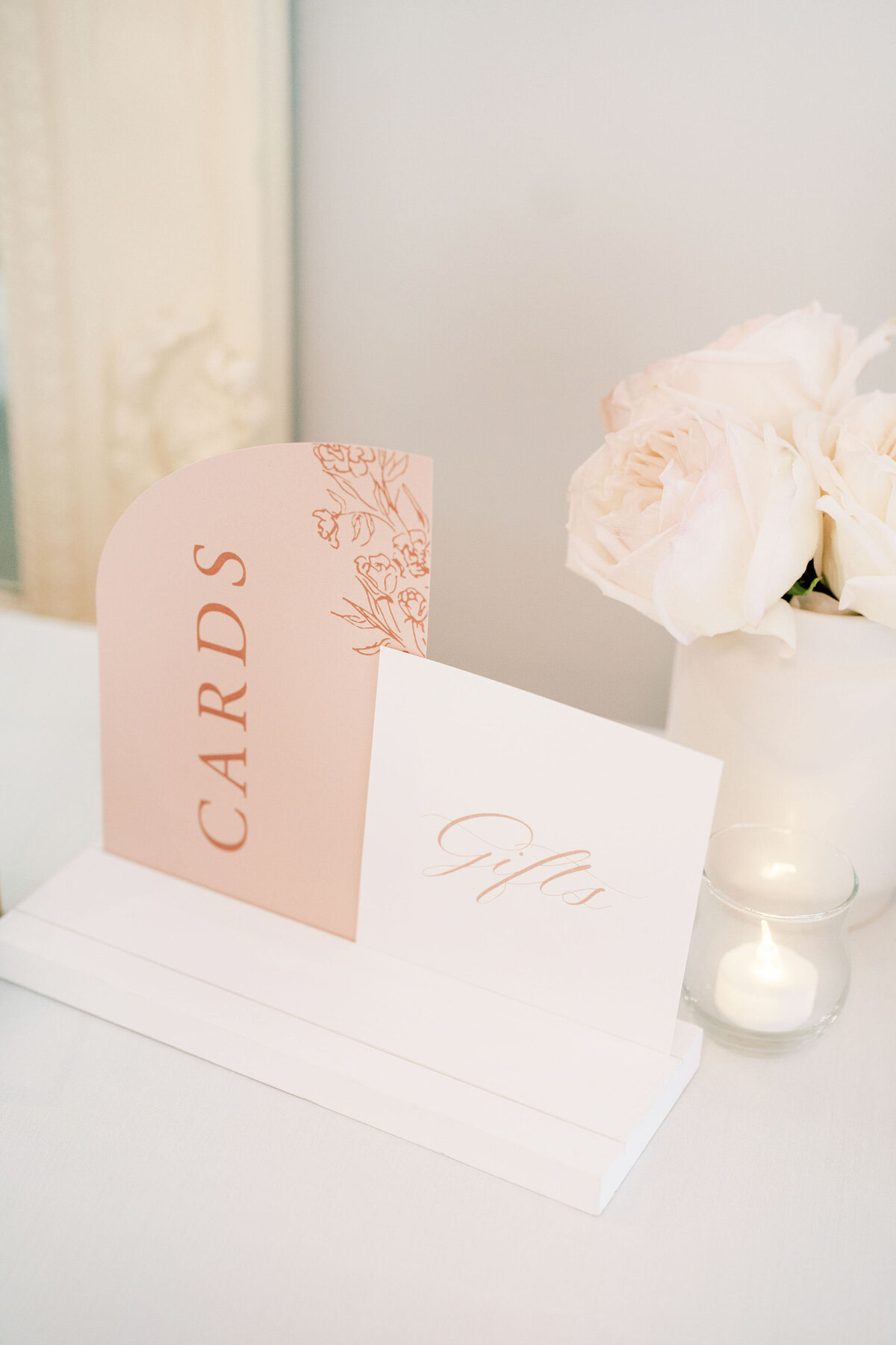 A table display at a Norland Estate wedding in Calgary features a stand holding two elegantly labeled cards, "CARDS" and "Gifts," alongside pale pink roses in a vase and a lit candle, creating a modern yet romantic ambiance.