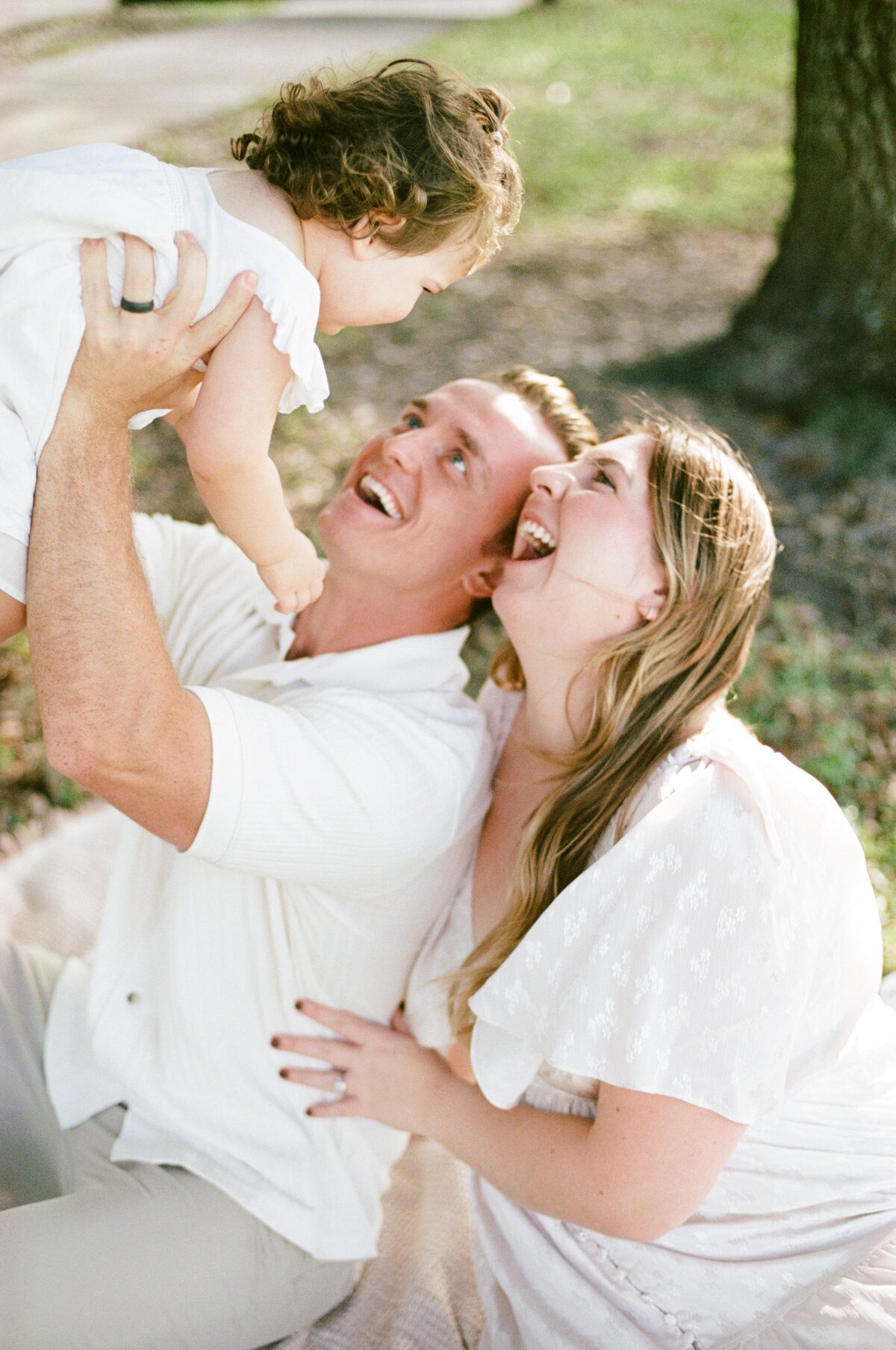 Family portrait taken at camp Milton in Jacksonville with mom and dad both wearing white and playing airplane with their toddler girl