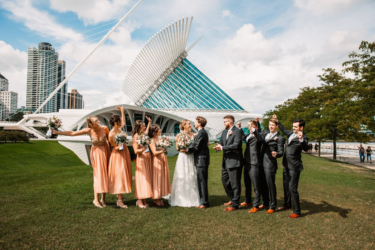 A wedding party cheers while the newlyweds hug in between them in the lawn of the Milwaukee Art Museum