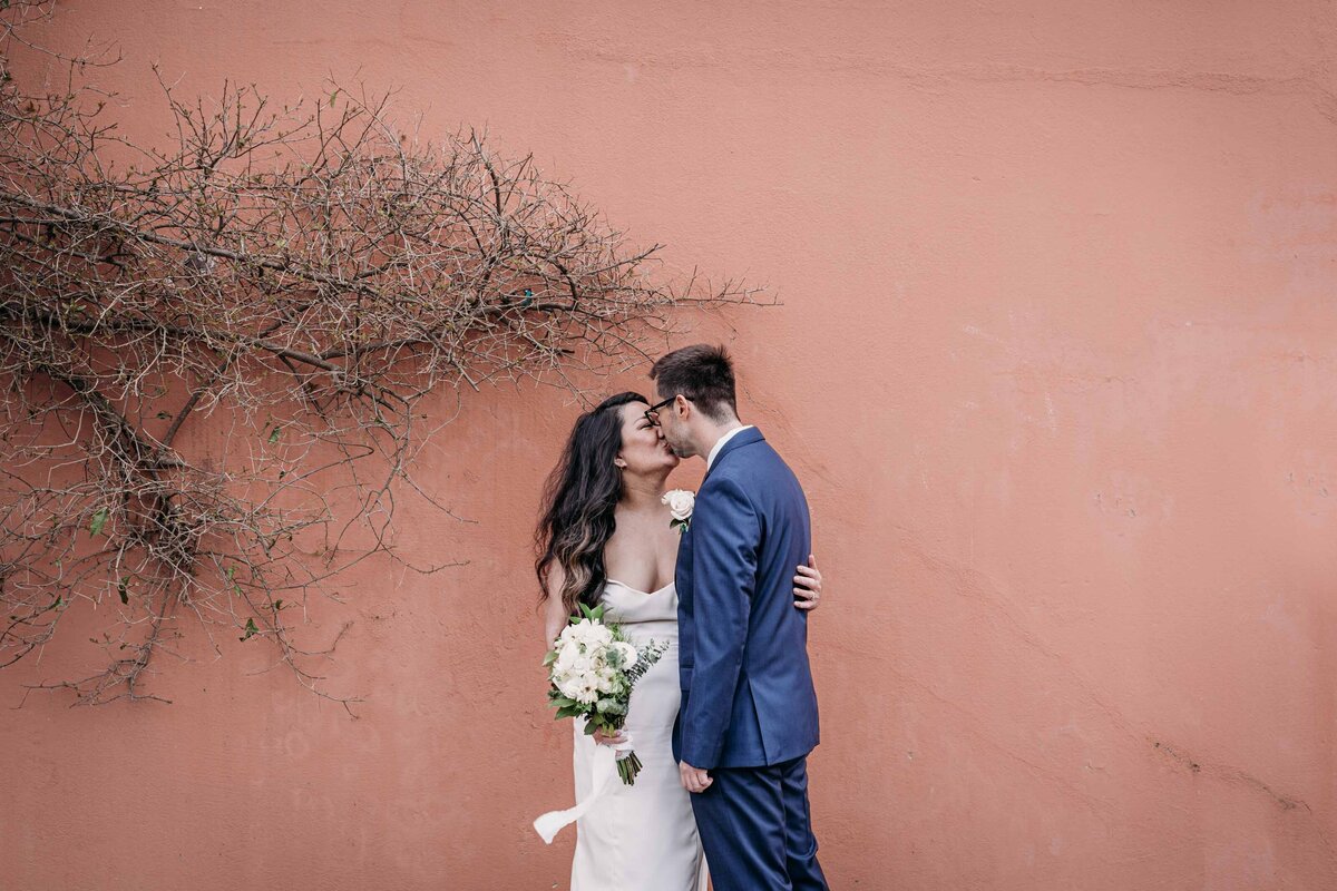 bold coral wall with dead vies on the left fram newly wed couple kissing on san francisco street.  Asian bride with long dark hair is wearing gown and holds a bouquet, white groom with glasses is waring a blue suit as he leans forward to kiss her.