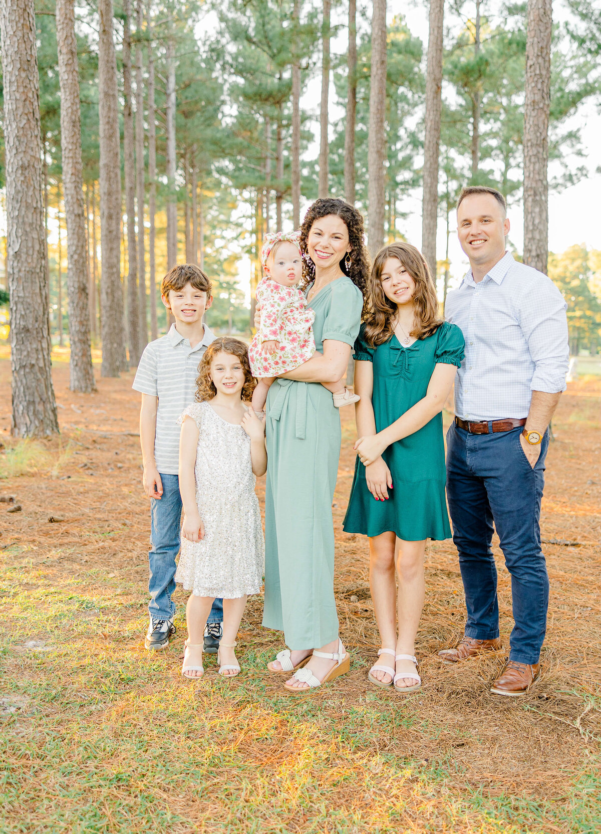 A family of six stands happily together in a forest, captured by a Fayetteville NC family photographer. The mother cradles a baby, while two girls and a boy stand nearby. The father smiles on the right as pine needles blanket the ground and tall trees frame this joyful moment.