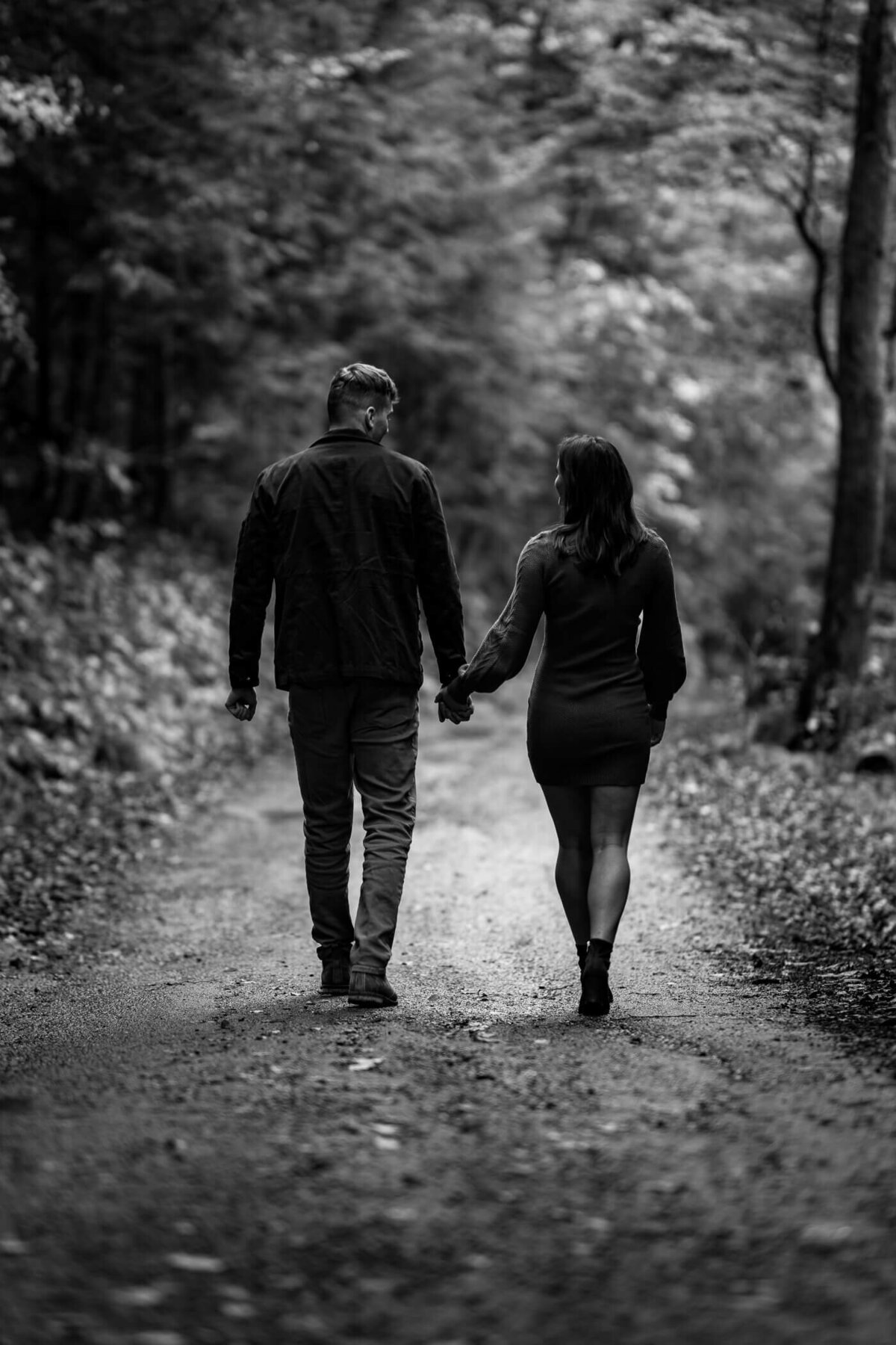 Black and White Pittsburgh engagement photography of a couple walking a holding hands down a dirt path. Captured during sunset at an engagement session at McConnell's Mill state park, PA