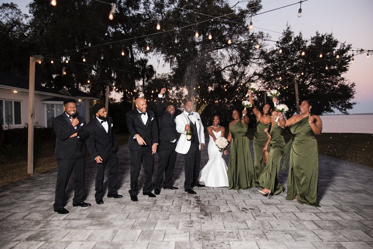 The bride and groom pop a bottle of champagne surrounded by their wedding party during a joyful celebration at Azaleana Manor in Orange Park, Florida. The groom wears a white tuxedo jacket while the bride, in a sleek white gown, stands beside bridesmaids in deep green dresses and groomsmen in black tuxedos. Under twinkle lights and mossy oak trees, this candid moment captures the excitement and fun of the evening. Documented by Heartful Films, a Jacksonville wedding photographer and videographer known for lively, true-to-color storytelling.