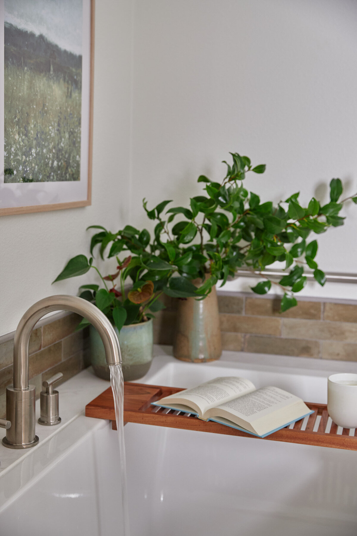 - Bathtub with silver arched deck mount faucet with water pouring out. Wooden bathtub tray with an open book and a white candle sitting on it. Two vases sitting on the edge of the bathtub with green plants in them. Artwork on the wall behind the bathtub in a natural oak frame with a painting of a field of flowers. - Bathtub is freestanding and wrapped in a white and grey marble. - Wooden stool with a white vase with green plant in it.