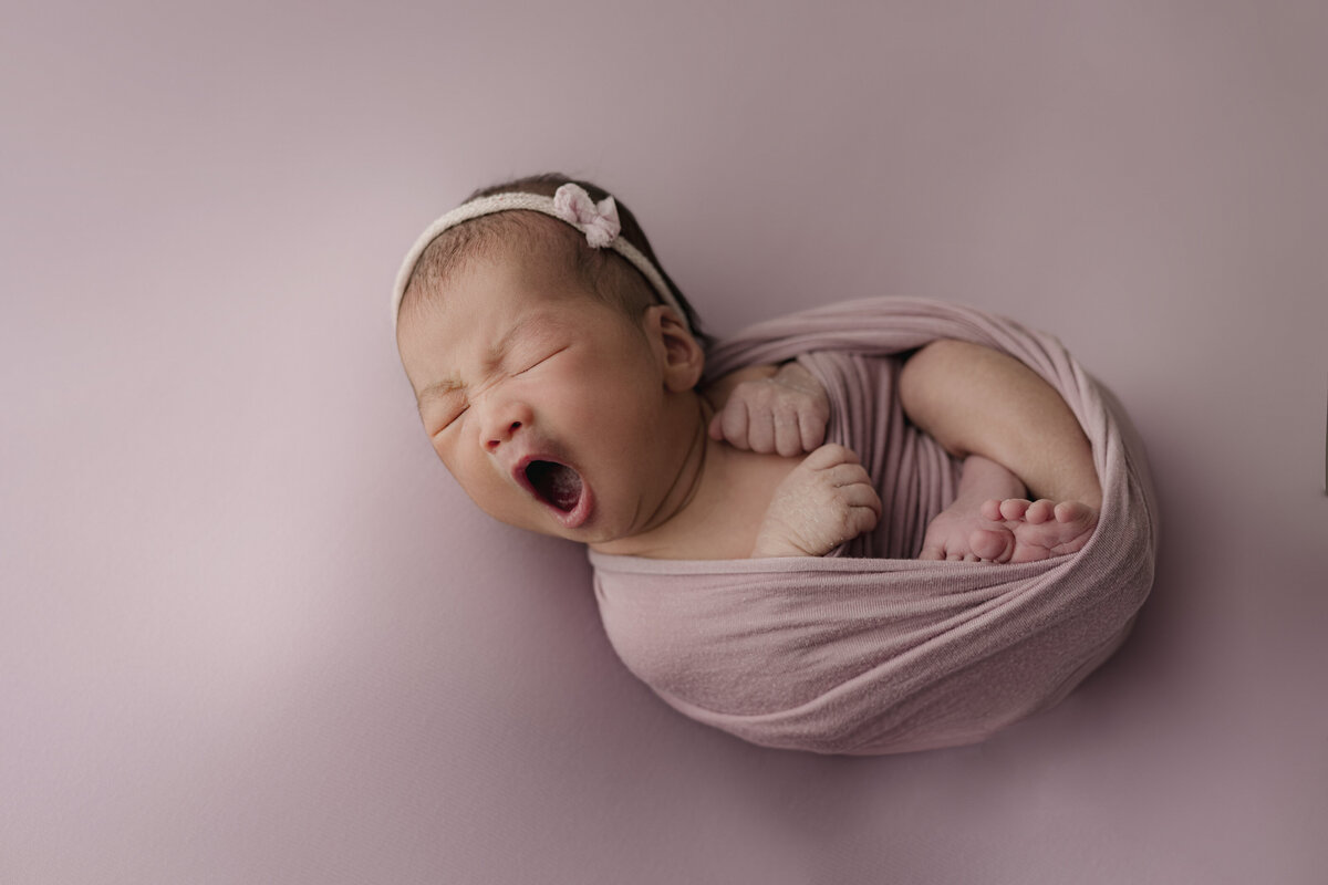 baby wrapped in pink yawning on pink backdrop
