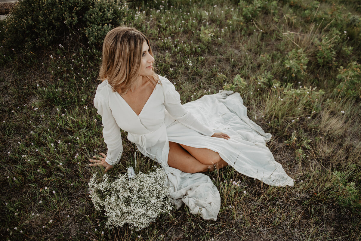 bride wearing a boho wedding dress with a high slit while sitting in a field and looking off into the distance