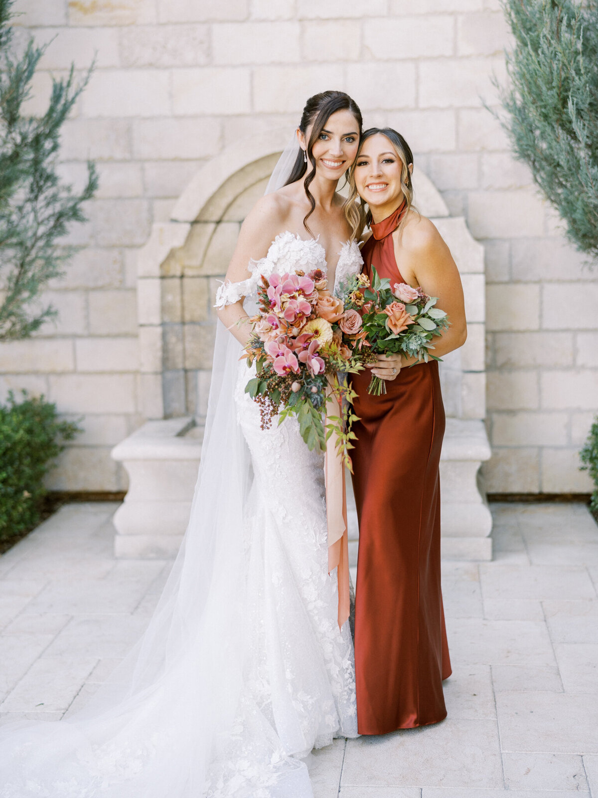 Bride and bridesmaid sharing a joyful moment in front of stone arches—timeless elegance with rich, romantic florals.