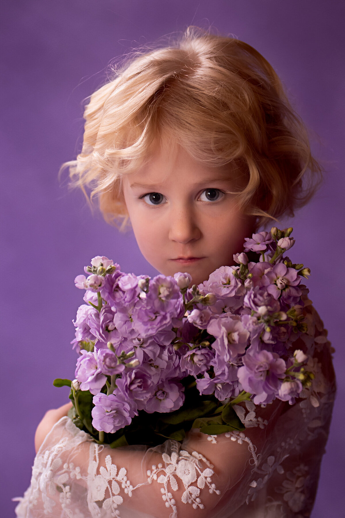 Delicate fine art portrait of a young child holding a bouquet of lilac flowers, captured against a soft pastel backdrop. Taken in Overland Park, Kansas, this setup is ideal for families seeking floral and artistic child photography.