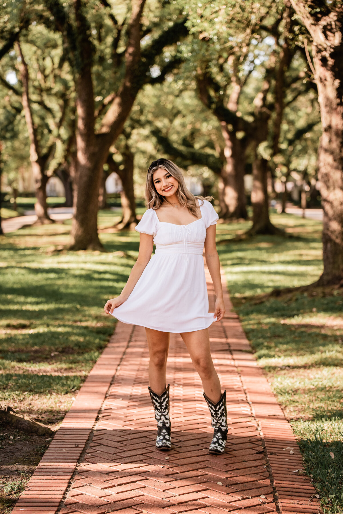 A senior holds the hem of her white dress and sways on a tree lined path.