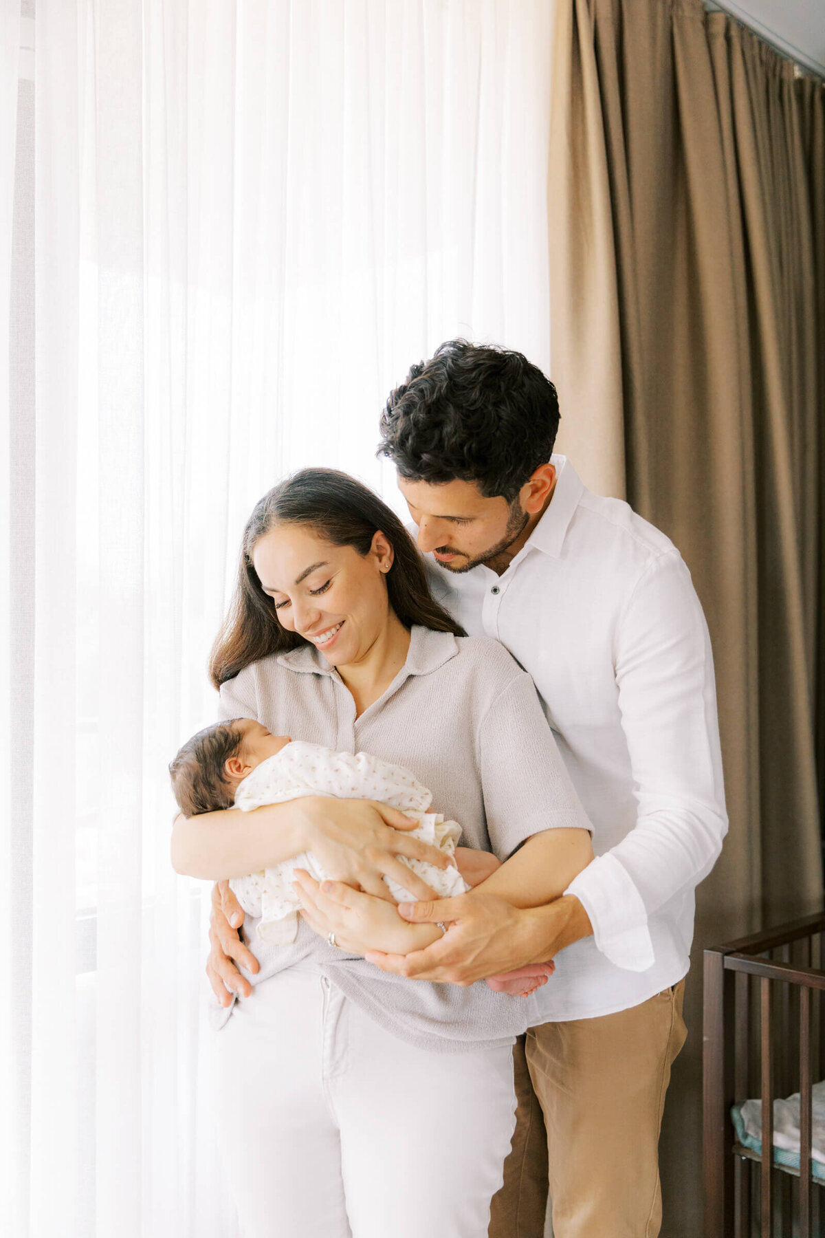 Een jong stel houdt hun slapende pasgeboren baby dicht tegen zich aan, vastgelegd in een rustige en lichte sfeer.