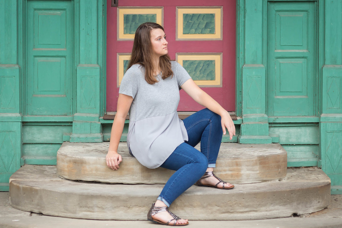 Waynedale High School girl sitting on steps of historic building in Zoar Village Ohio, building door is painted burgandy and yellow with a bright green background on building, senior girl is not looking at camera but off to side, she is sitting turned to the left, with one leg up higher on one step, photographed by Jamie Lynette Photography Canton Senior Photographer