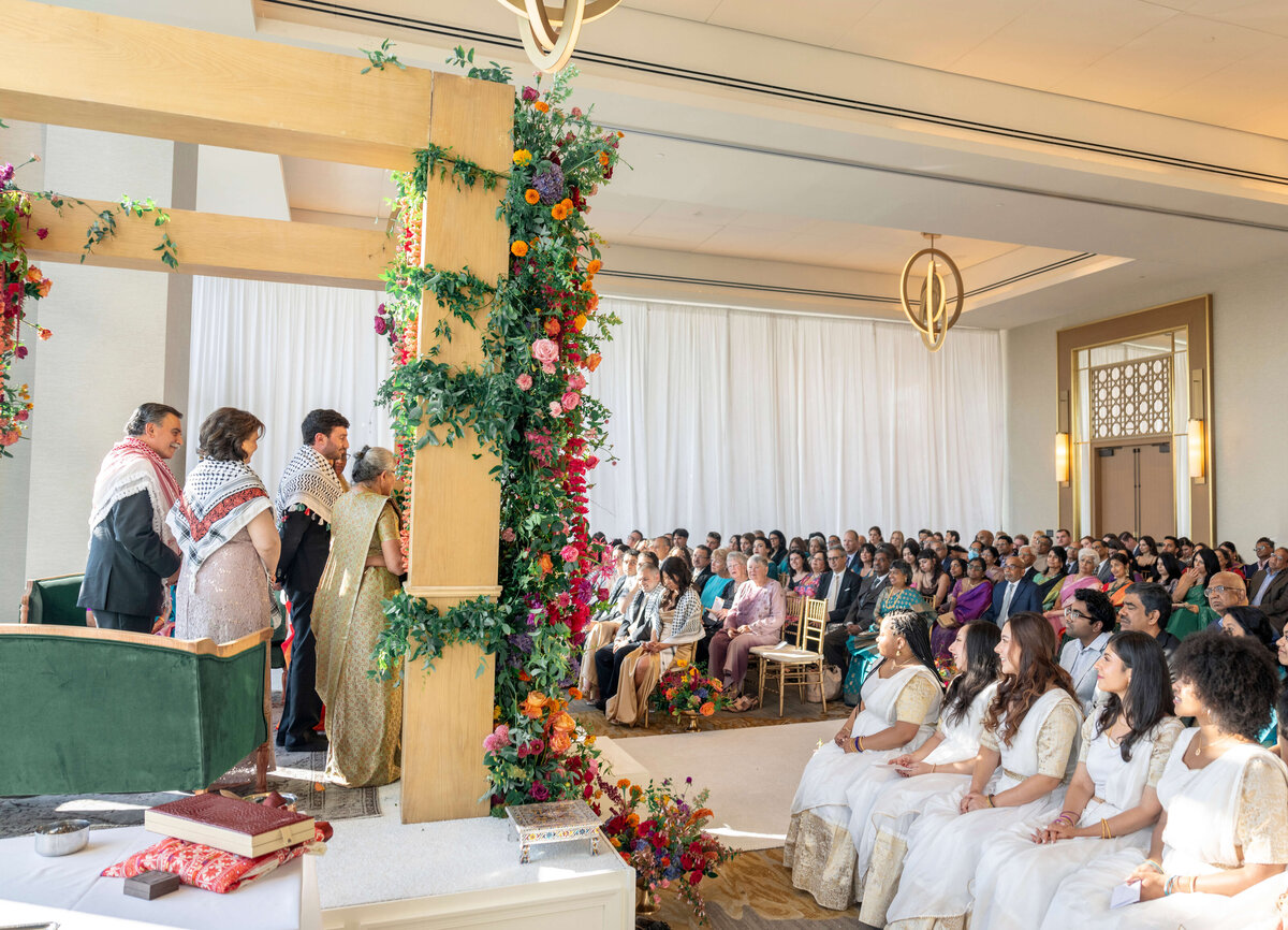 A wedding ceremony takes place indoors under a floral arch with vibrant flowers. A couple stands with several people, possibly family, on the left. Guests sit attentively on the right, dressed in formal attire, some in white and traditional outfits.