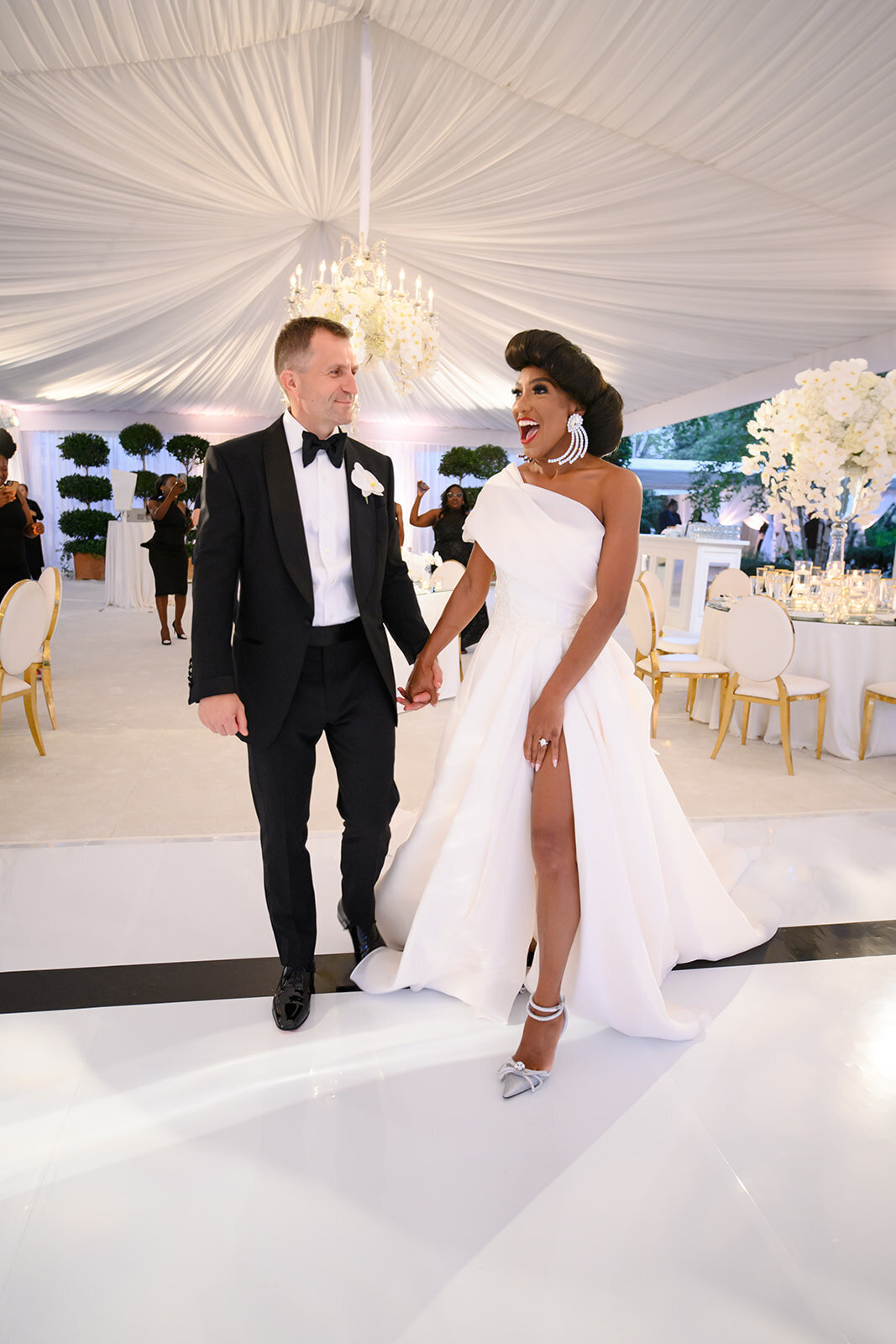 Bride and groom dancing exuberantly in a black and white photo, surrounded by a floral backdrop and guests at their reception.