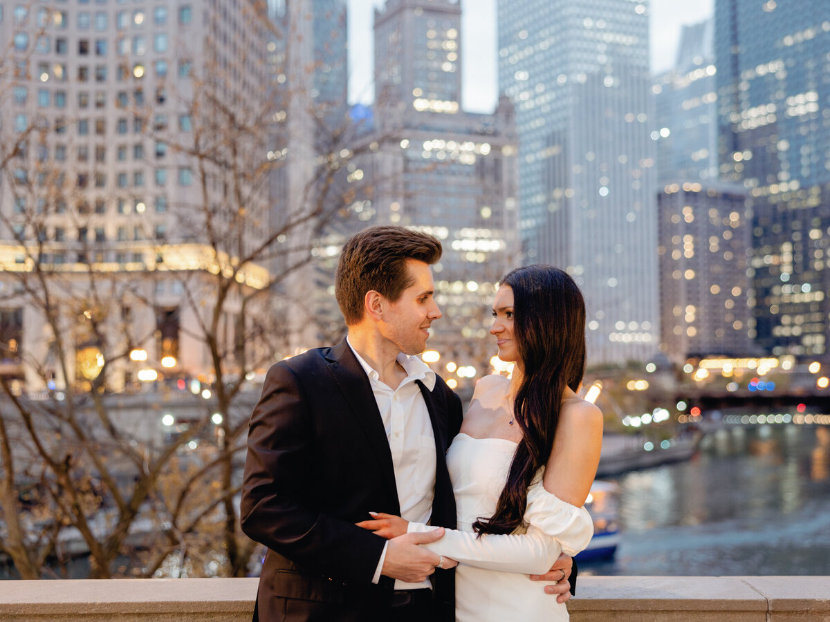 A couple embraces at dusk along Michigan Avenue and the Chicago River, with the city lights creating a romantic and timeless backdrop.