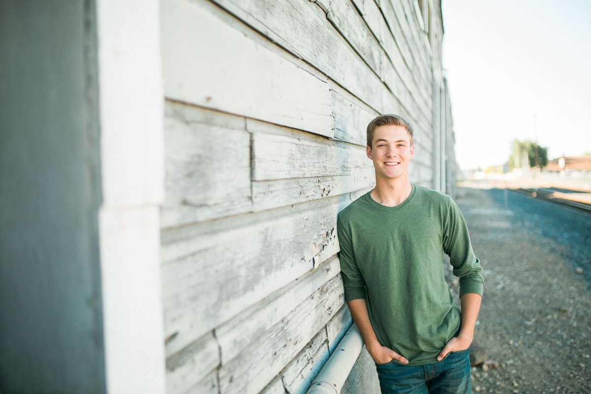 Senior male poses for his portraits leaning against a wall next to train tracks