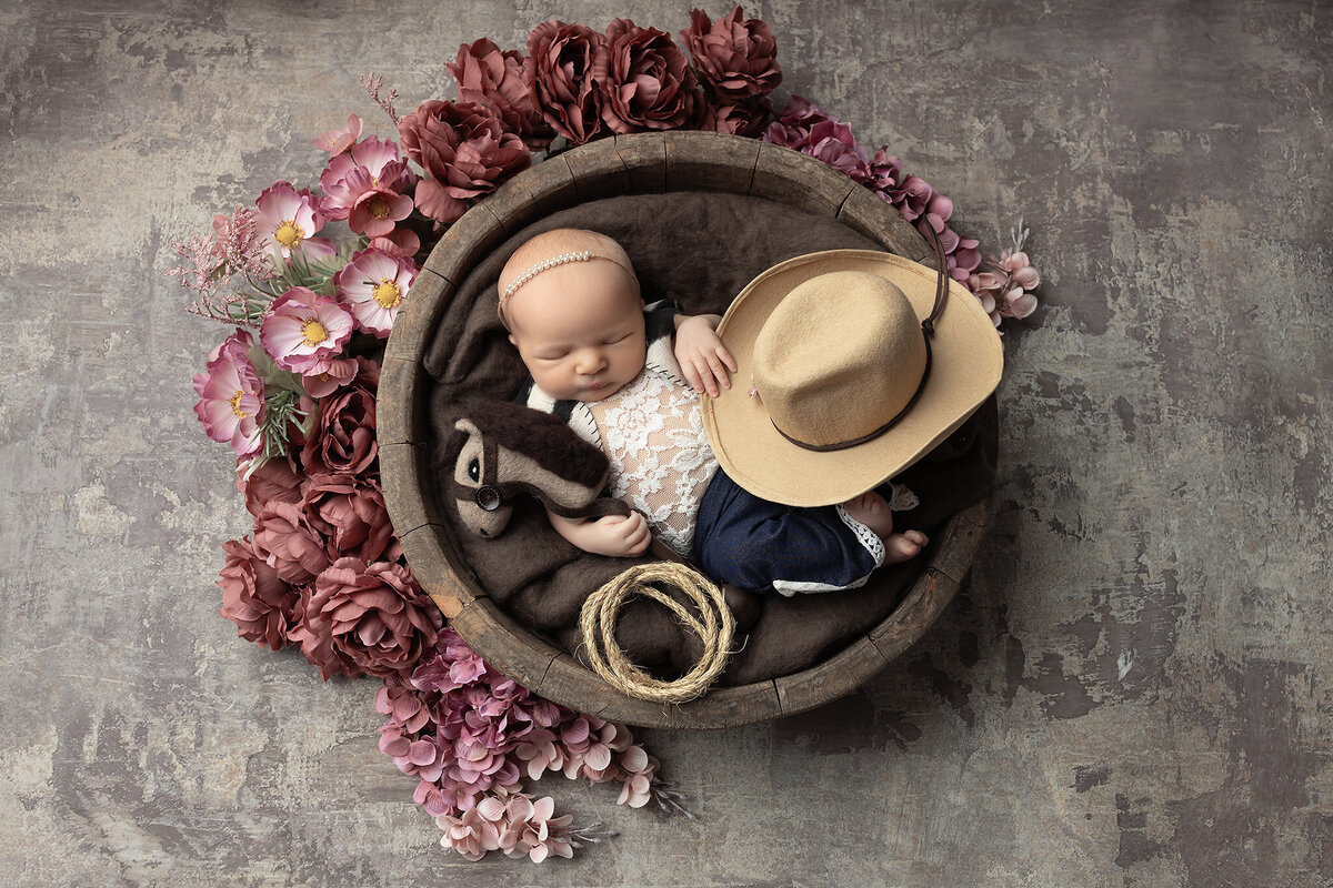 Newborn girl in cowgirl outfit with a hat, horse toy, and rope, styled with blush flowers for western baby photography by a DFW newborn photographer.