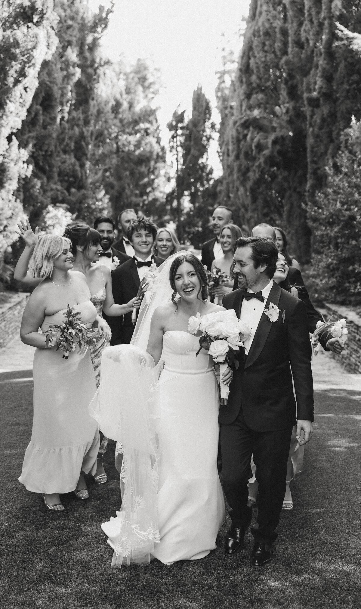 bride and groom walking and laughing with bridal party walking behind them  in black and white