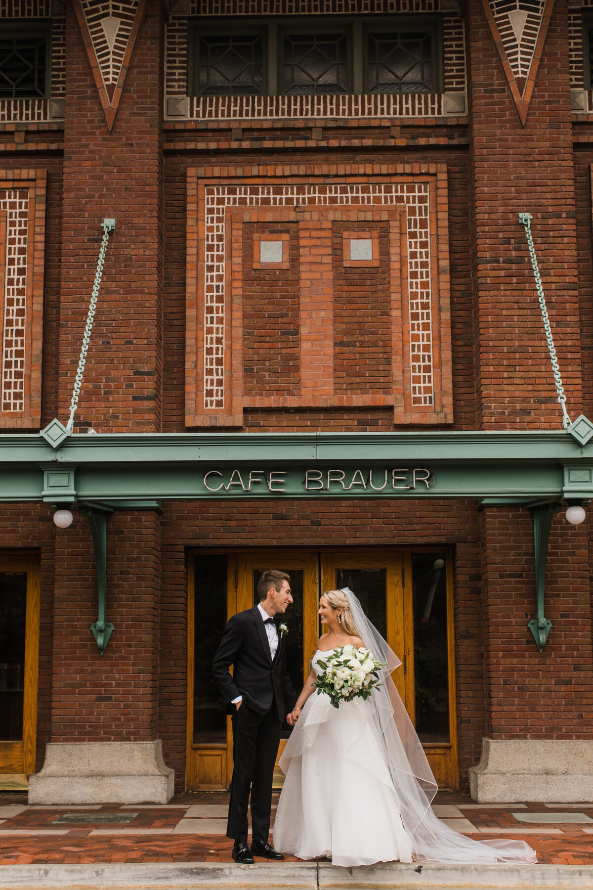 A bride and groom pose in front of the Cafe Brauer entrance for a wedding photo