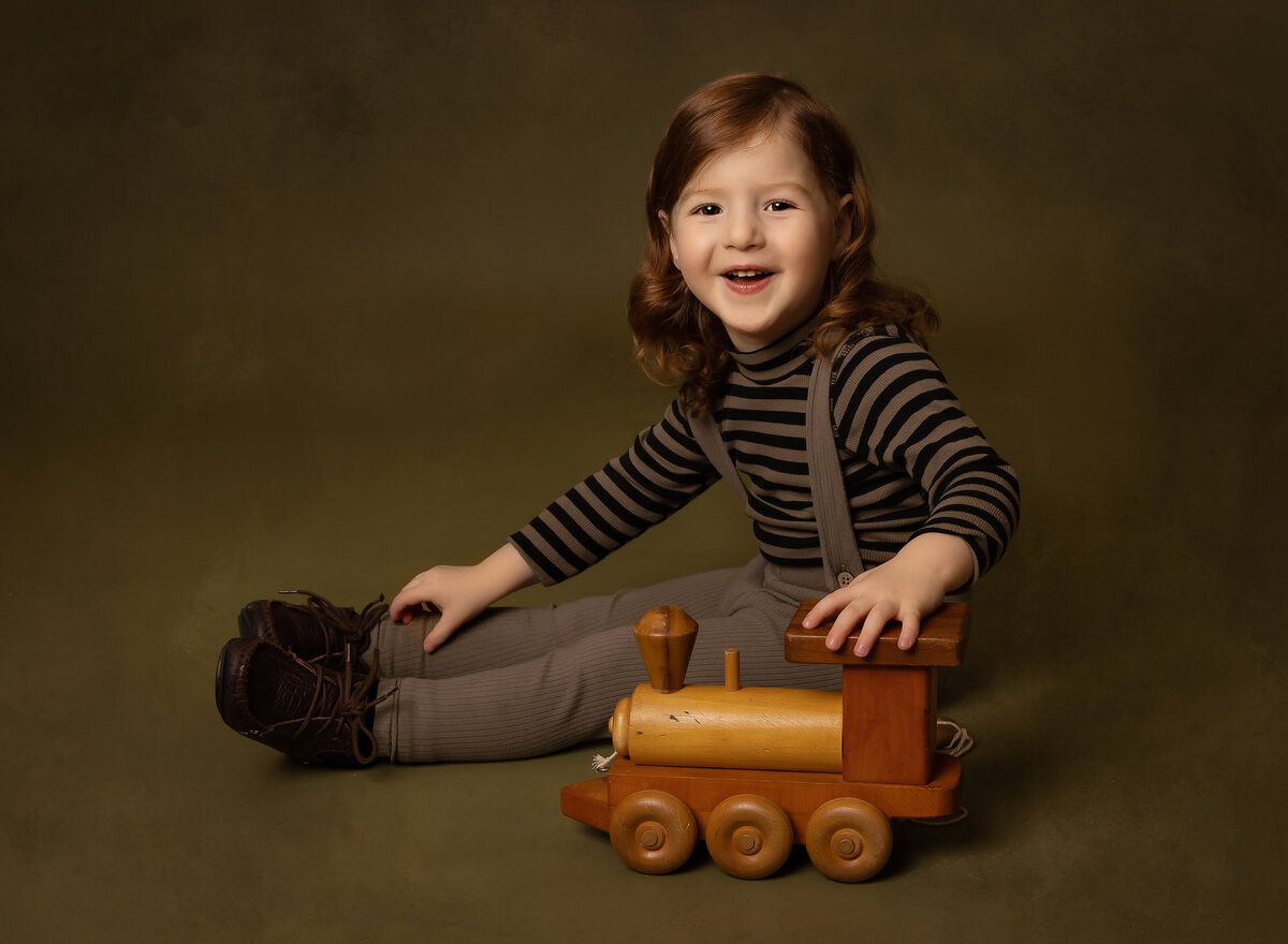 Little boy sits on the ground with a wooden vintage train for a portrait to commemorate his upsherin. He is looking over his shoulder and smiling at the camera. Captured by brooklyn family photographer Chaya Bornstein.