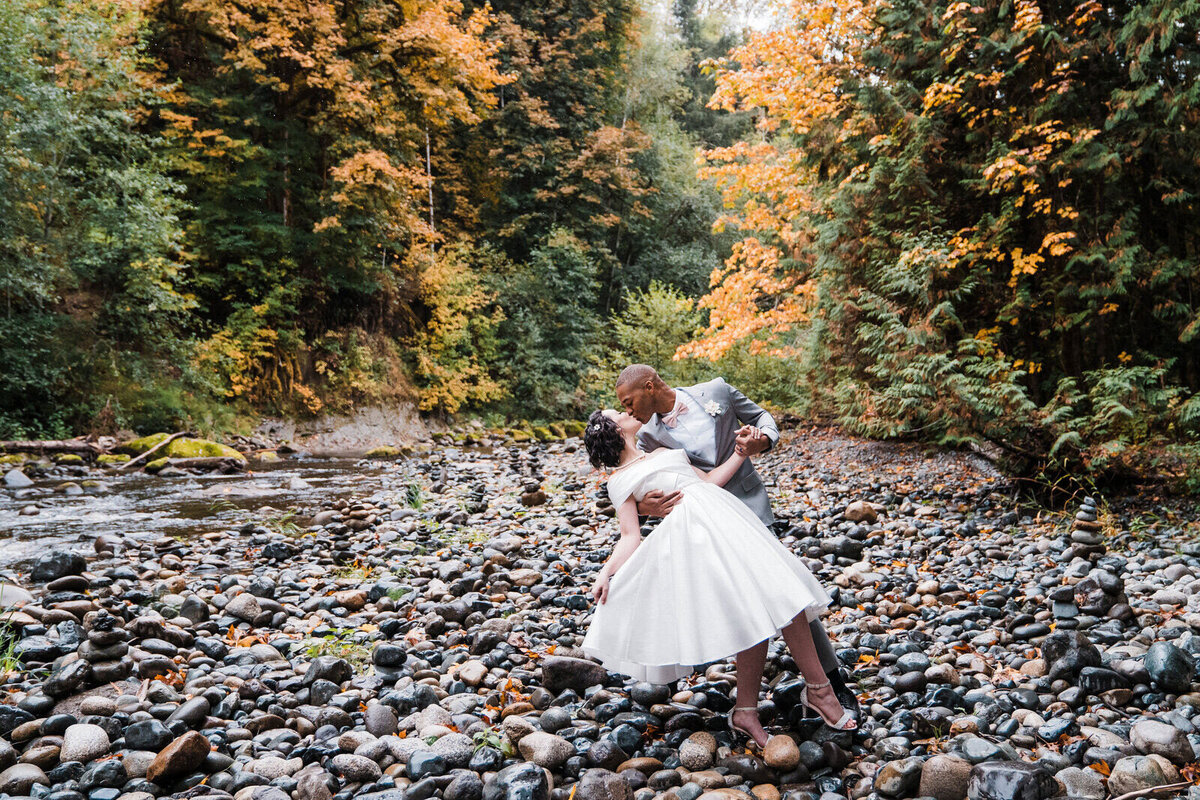 A groom dips his bride while standing on river rocks surrounded by autumn colors in the Washington cascade mountains