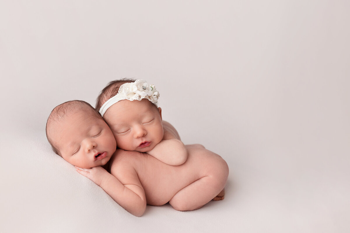 Newborn twin babies posed together on a cream backdrop in a side-lying snuggle pose wearing matching headbands.