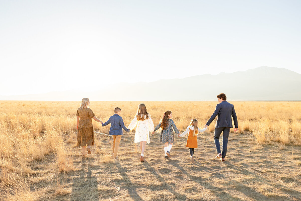 A family of six walks away from the camera, holding hands as they stroll through a golden field at sunset in Eagle Mountain, Utah.
