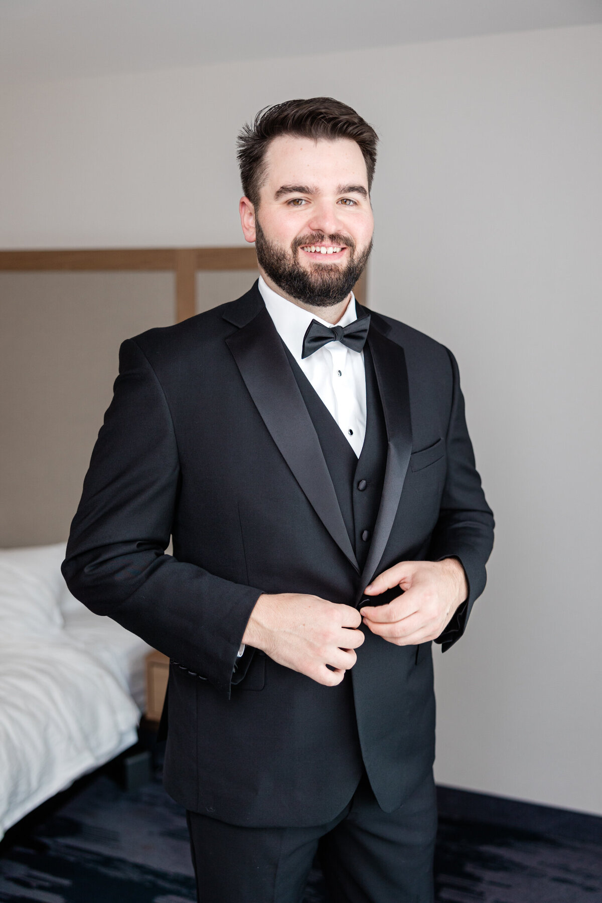 A groom smiles at the camera as he  buttons his tuxedo