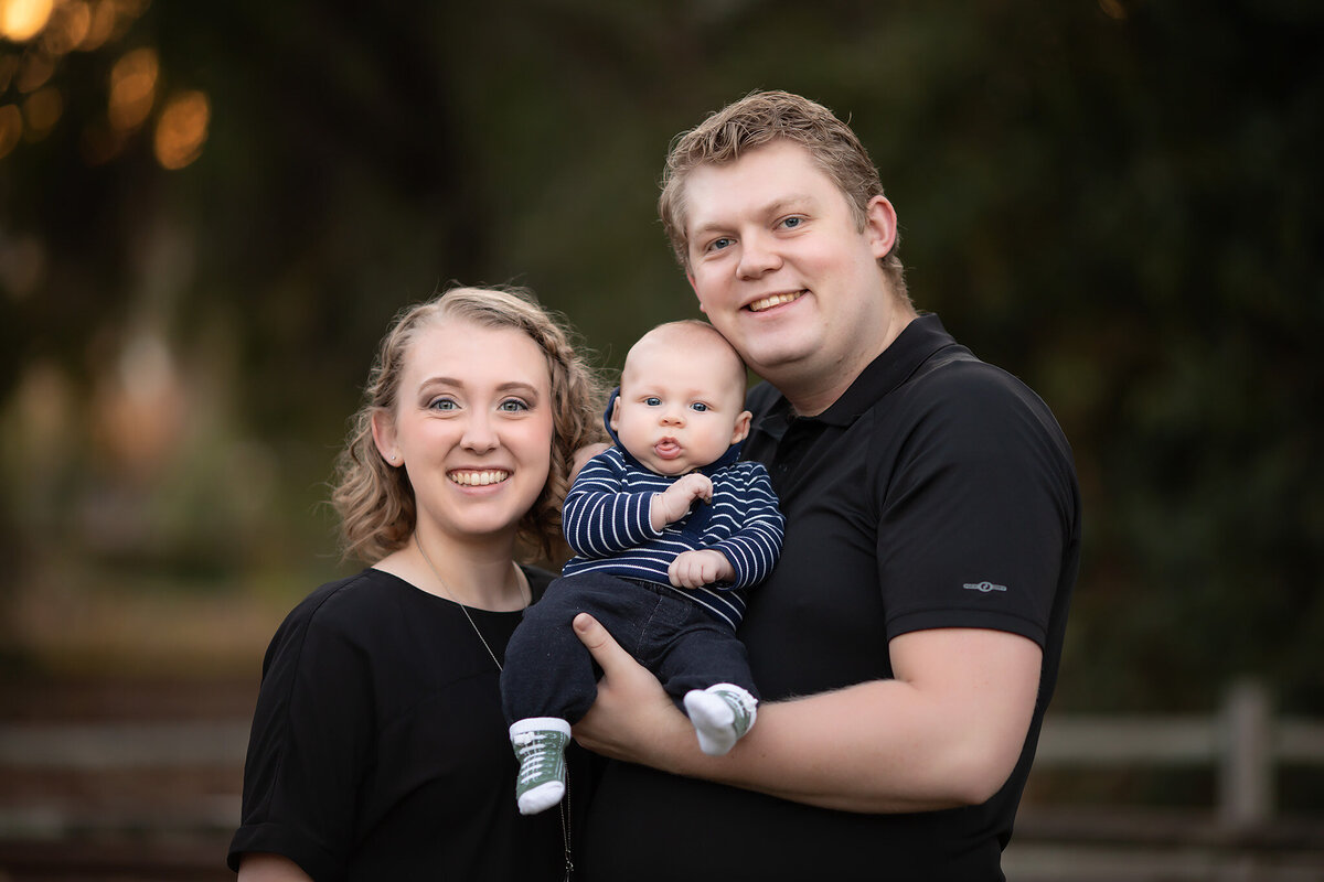A happy family with a baby in their arms, smiling together in a park setting, captured by a Dallas family photographer.