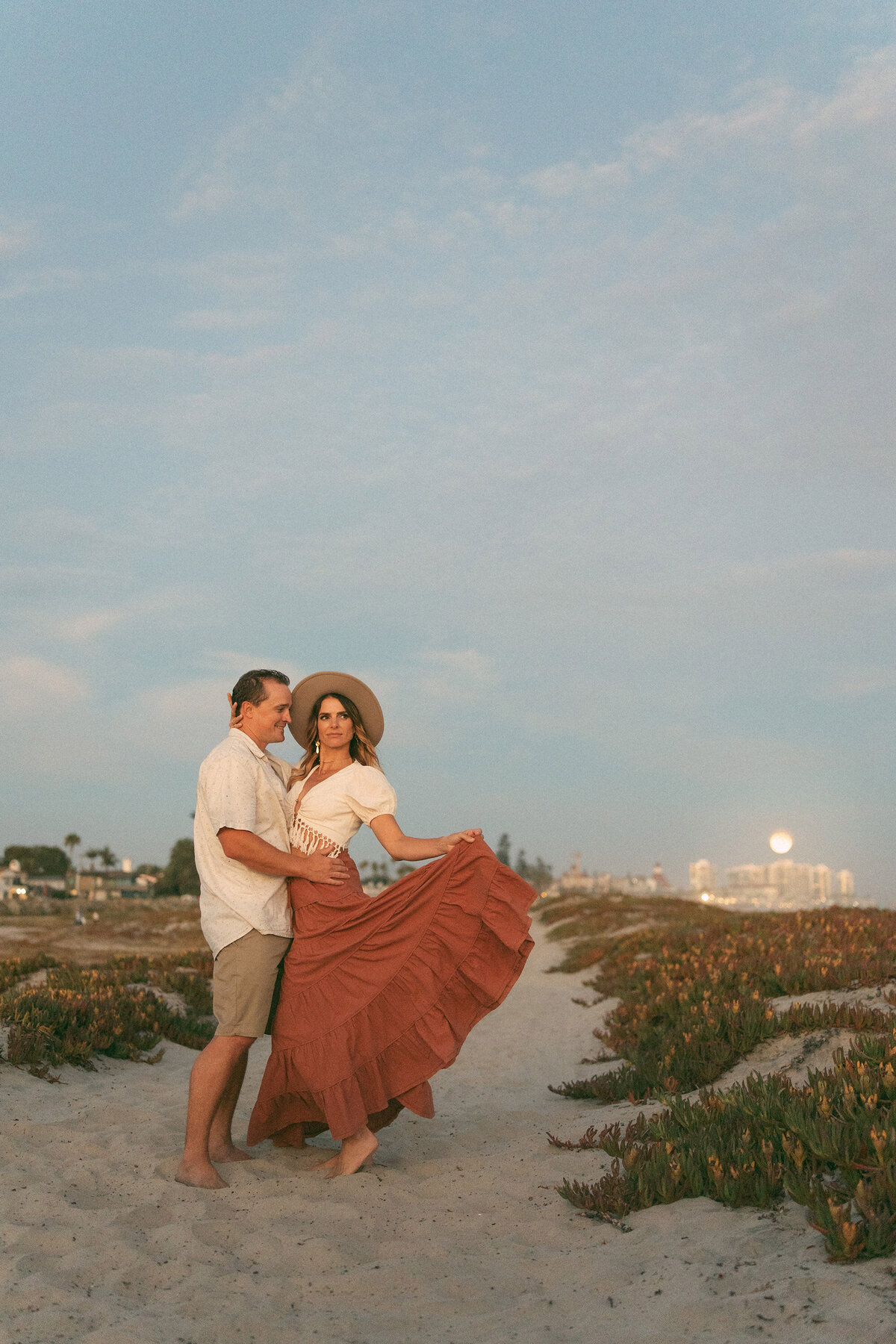 Romantic couple sunset session on the beach in Coronado, San Diego – barefoot partners embracing on sandy path with the woman in a flowing rust-colored skirt and wide-brim hat, golden light and city skyline in the background