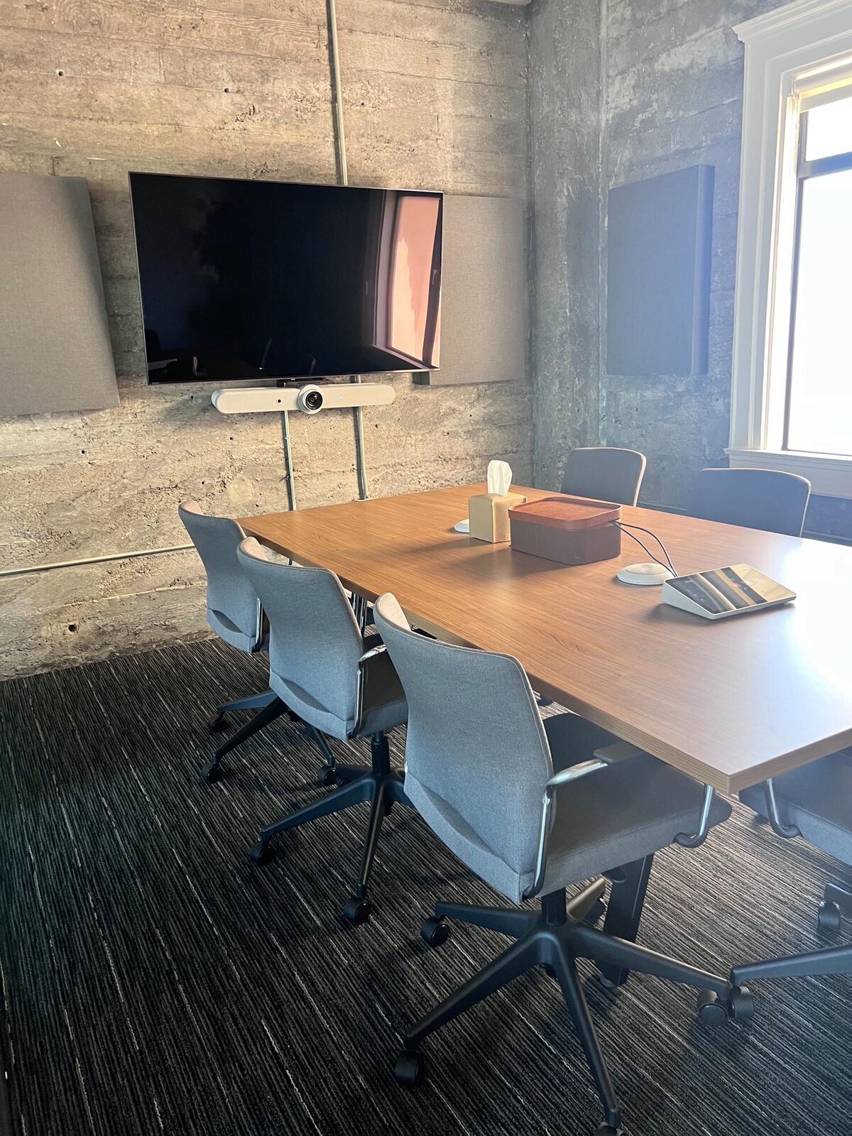 Small conference room with a wood meeting table, gray chairs, wall-mounted TV, and acoustic panels against a concrete wall.