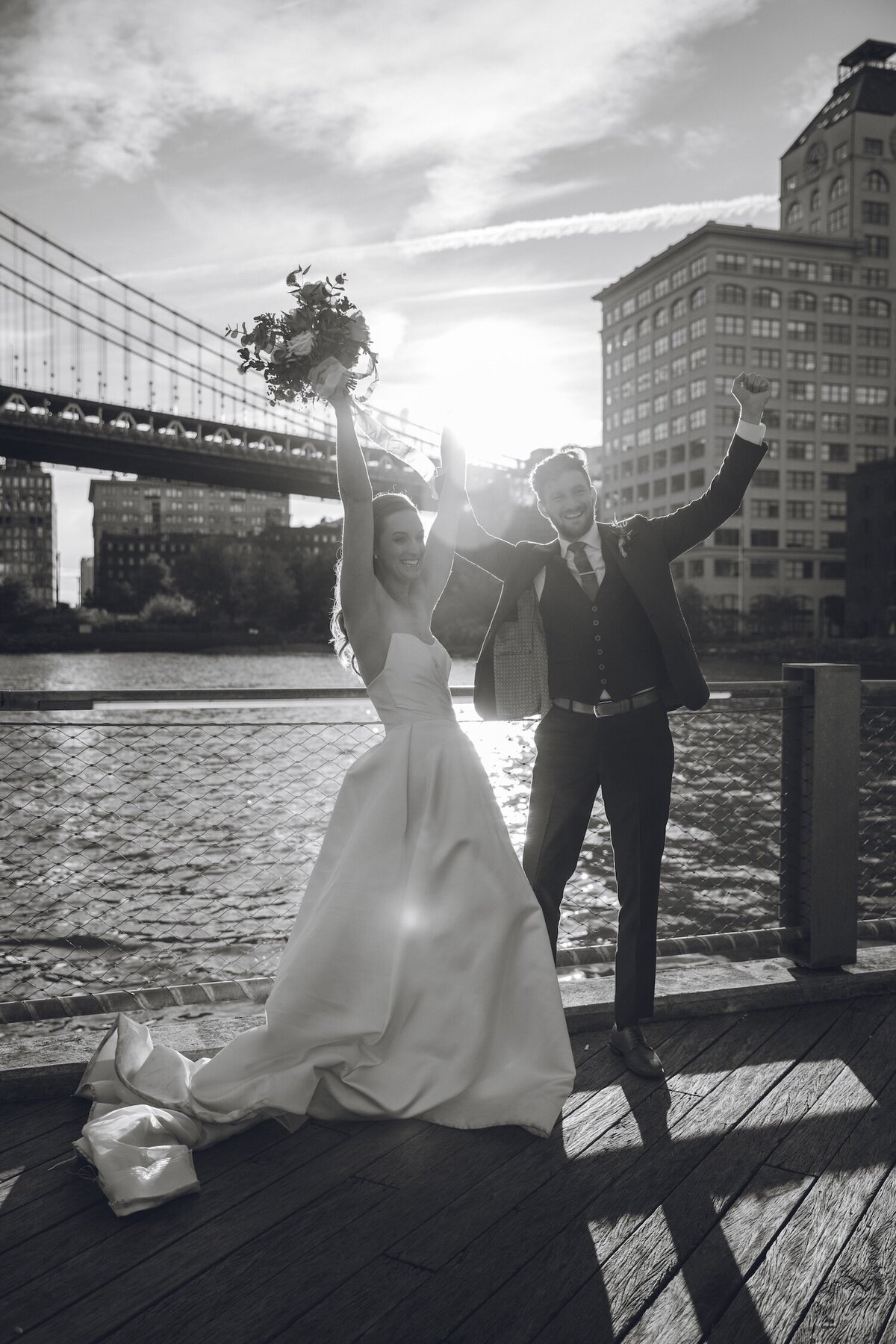Bride and groom cheering with bouquet at Brooklyn Bridge Park