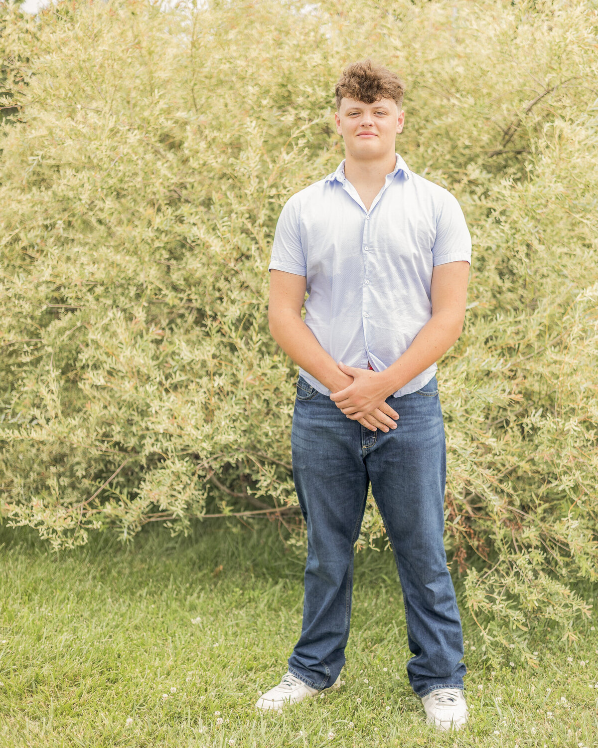 High school son is standing in the grass in front of a bush with his over one another looking at the camera with a soft smile.  He has short light brown hair, wearing a light blue, short sleeve button up shirt with blue jeans.