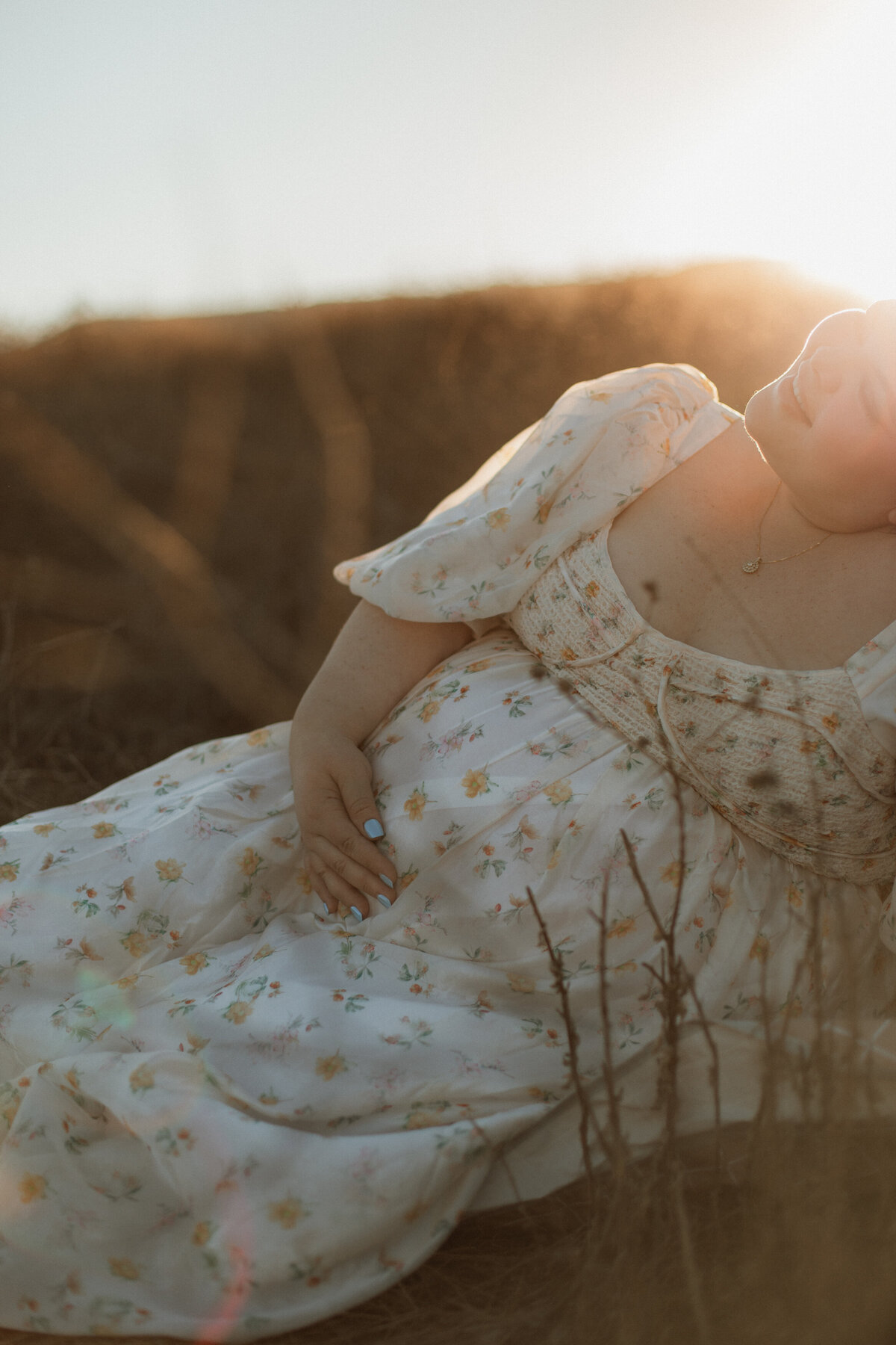 A pregnant woman sitting in a sunlit field, smiling as warm sunlight glows behind her, hand resting on her floral dress-covered belly.