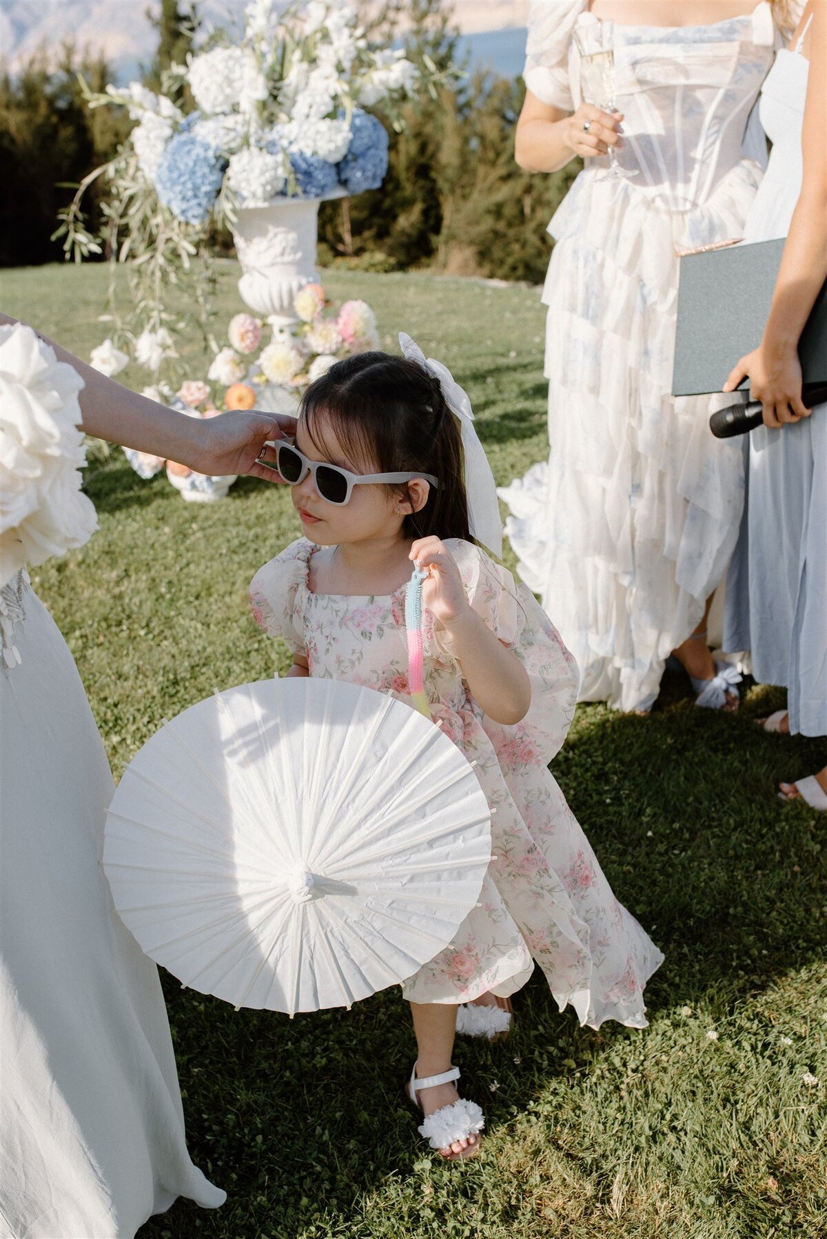 Flower girl with parasol and sunglasses receiving a gentle touch while standing near floral ceremony installations at a Lake Hāwea wedding.