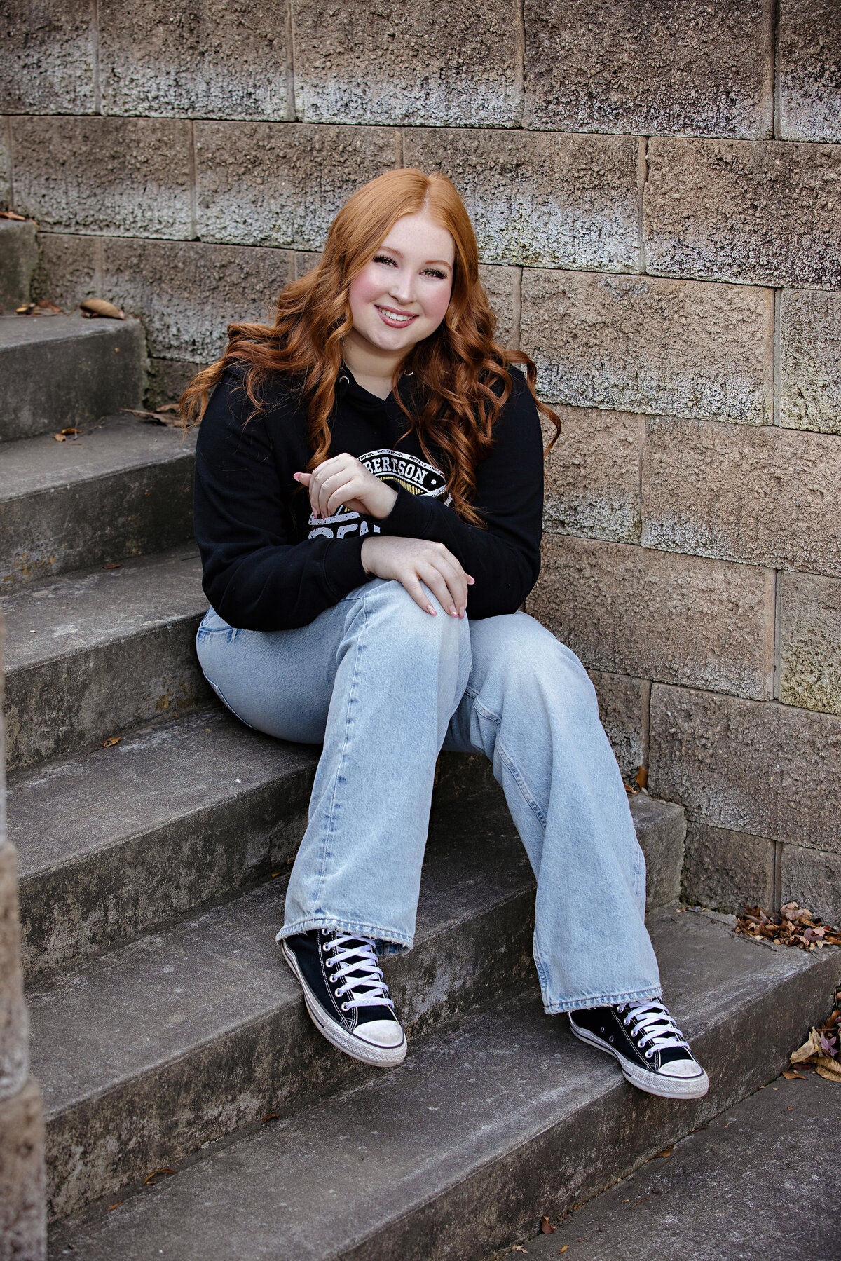 High school senior in black hoodie sitting on concrete steps with relaxed pose 