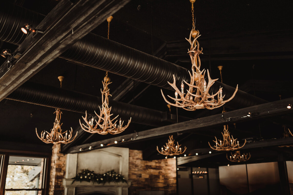 A warm and intimate wedding reception at The Lake House in Calgary, viewed from outside through large glass windows. Guests are seated at long, candlelit tables, engaged in conversation and enjoying the elegant atmosphere. The rustic charm of the venue is highlighted by stone walls, a grand fireplace adorned with greenery and candles, and antler chandeliers casting a soft glow. The dim lighting and reflections in the glass add depth to the scene, creating a cozy and romantic ambiance for the celebration.