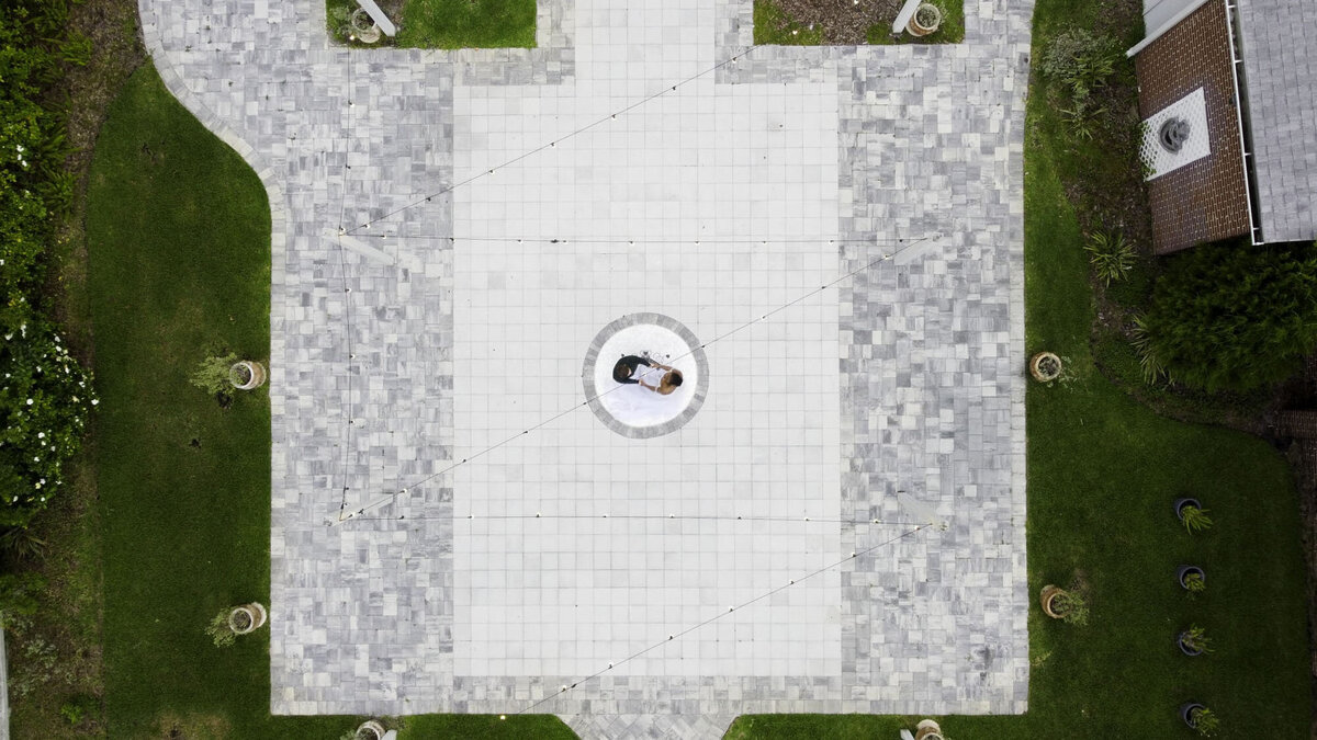 A stunning overhead drone view captures a bride and groom lying together at the center of a circular design on the stone courtyard of 5 Points Chapel in Jacksonville, Florida. Surrounded by a symmetrical layout of gray and white pavers and lush green grass, the couple shares an intimate, artistic moment. Captured by Heartful Films, a Jacksonville wedding photographer and videographer known for blending cinematic creativity with meaningful storytelling.