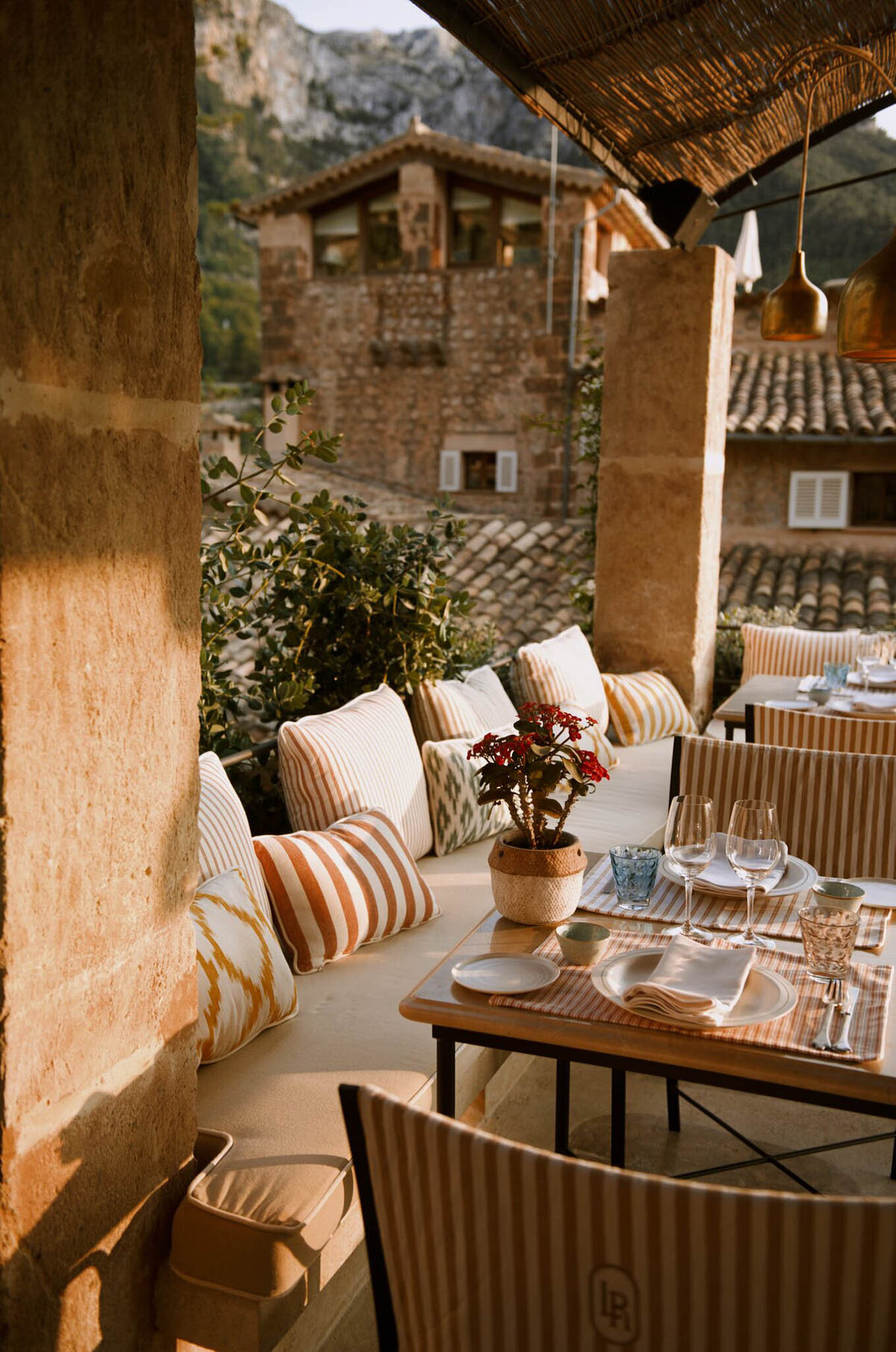 Outdoor dining area with cushioned seating and striped pillows on a stone terrace. A set table with wine glasses and a potted plant centerpiece overlooks rustic buildings with tiled roofs and a mountainous backdrop. Warm sunlight casts soft shadows.