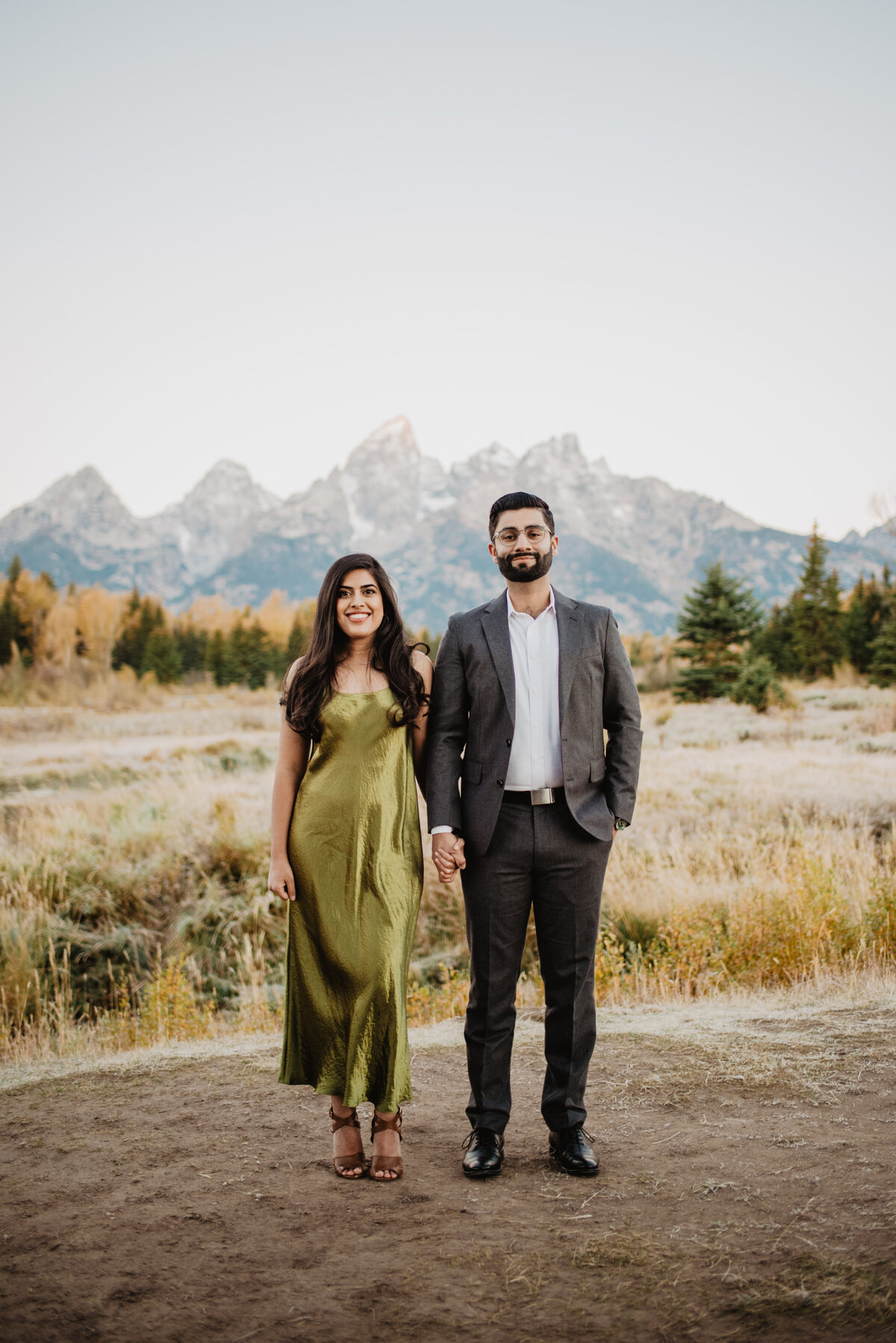 photographers in jackson hole photographs formal engagement session in Jackson Hole during the fall with man and woman holding hands in front of the Gran dTetons, man in suit and woman in velvet dress