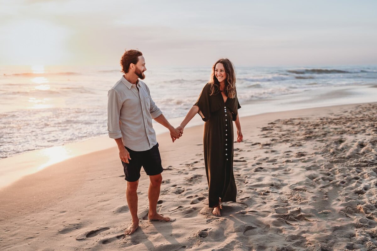 Couple holding hands and walking barefoot along the beach at sunset during a San Diego lifestyle photography session.