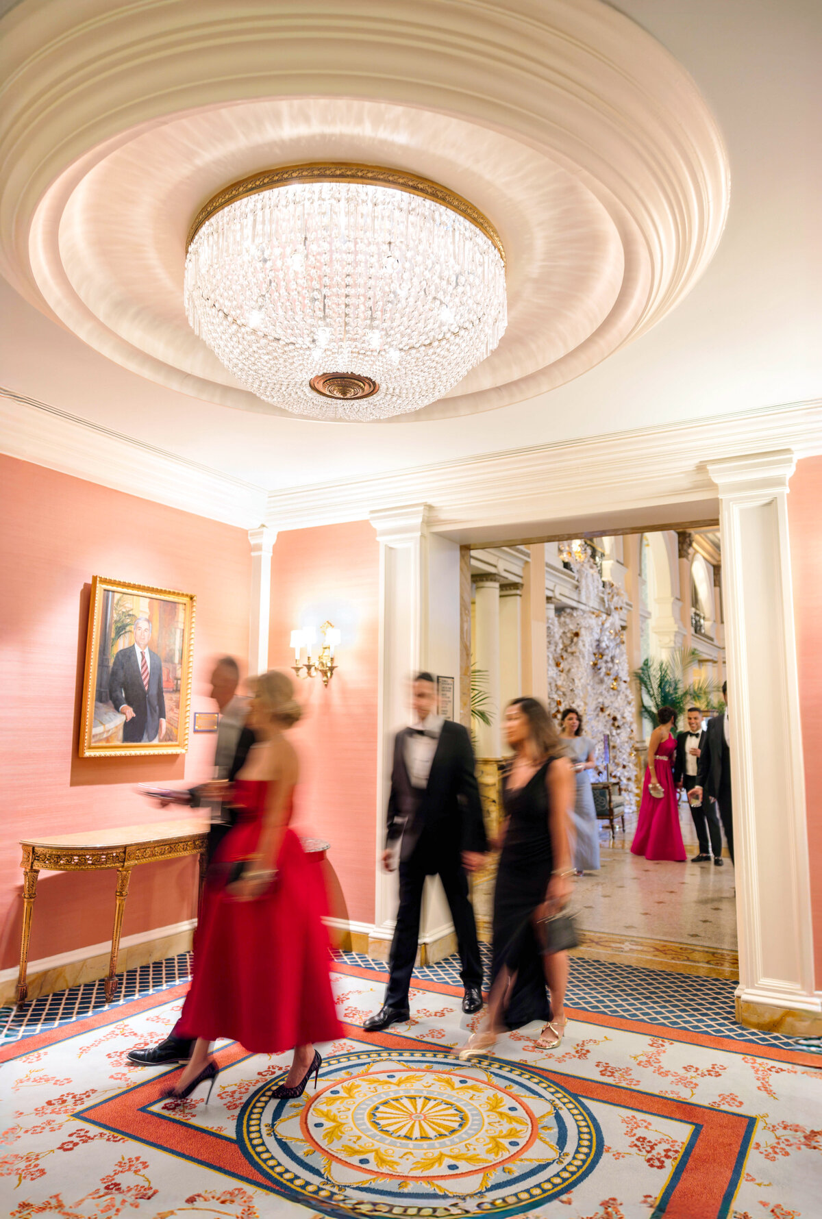 People in formal attire walk through an elegantly decorated hallway with a chandelier and a portrait on the wall. The floor has an intricate pattern, and more individuals are visible in the background, suggesting a bustling, upscale event.