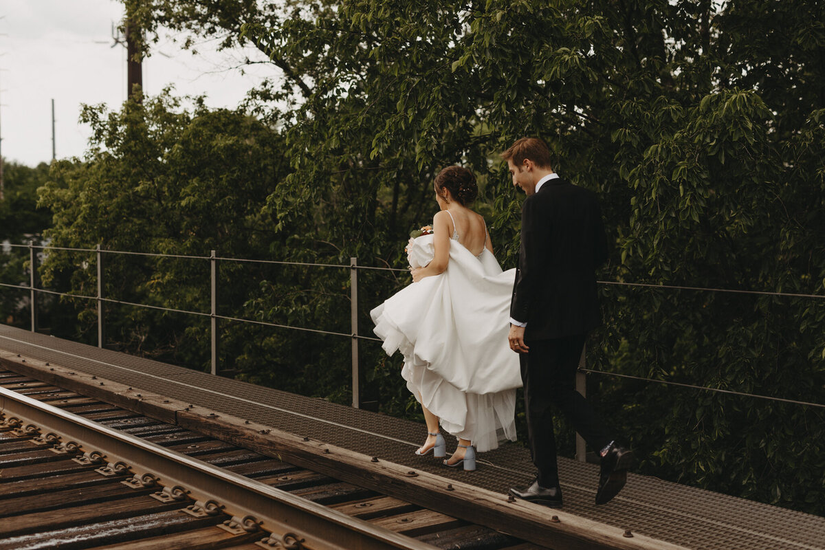 groom helps bride carry dress as they walk on the side of a railroad track