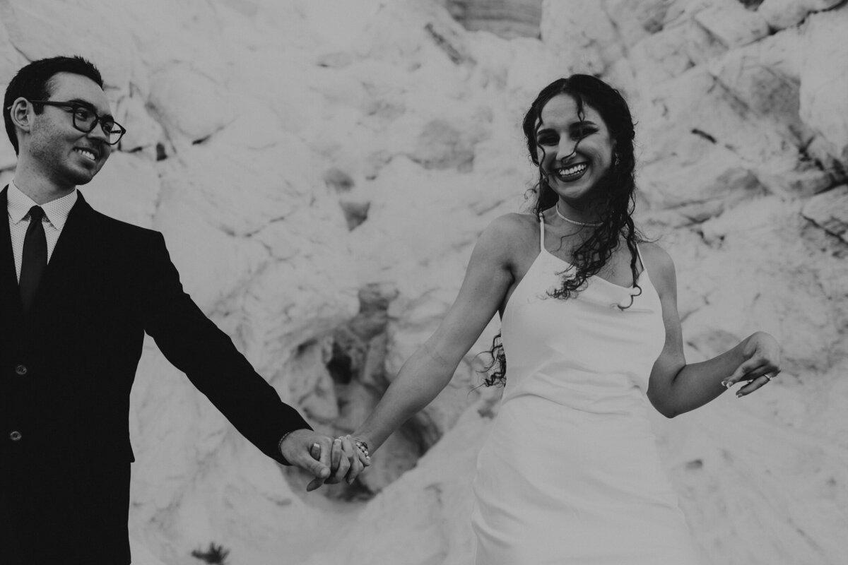 bride and groom walking while holding hands in front of a white rock