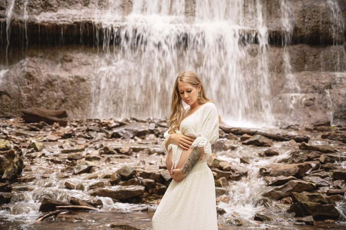 Woman expecting cradles her belly in a gorgeous water falls scenery behind her.