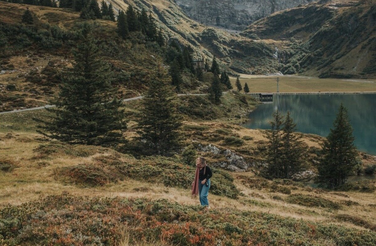 girl walking through the switzerland alps far away. She is looking up the hill with a camera around her shoulder. She has on a pink scarf and is walking up hill. There are greens and reds and yellows in the grass surrounding her.